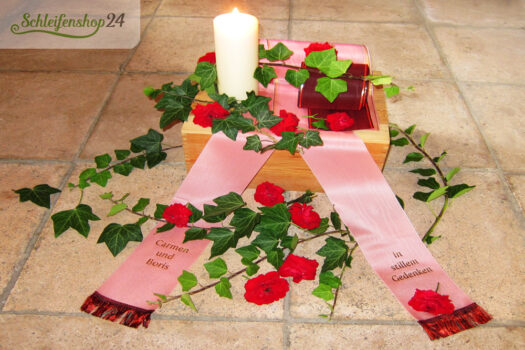 Trauerschleife in der Farbe pfingstrose liegt mit Efeu und einer Kerze auf einem Steinboden