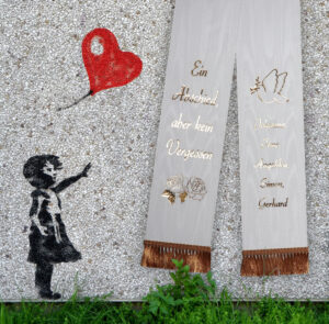 Der Trauerspruch Ein Abschied aber kein Vergessen auf einer Schleife vor einer Steinwand mit einem Mädchen und einem Luftballonherzen