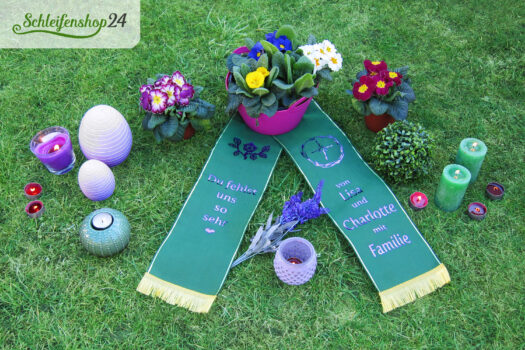 grüne Trauerschleife mit Text und Ornamenten in lila auf Gras liegend